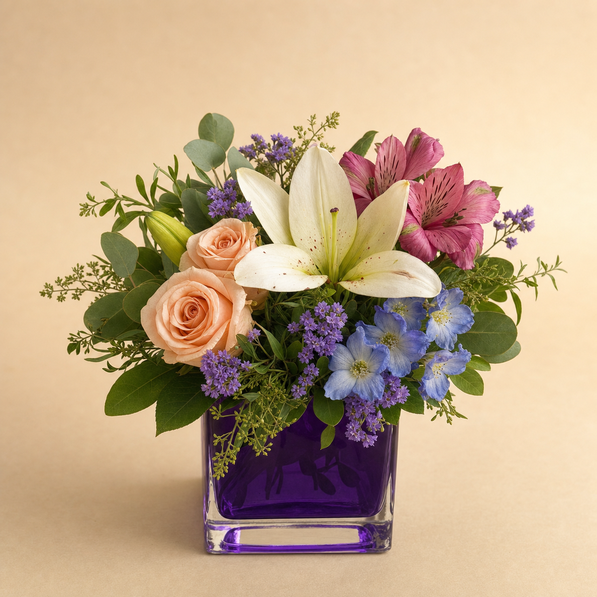Enchanted Blossoms Standard – compact arrangement of white asiatic lilies, peach spray roses, pink-mauve alstroemeria, blue delphinium and lavender statice in a purple cube vase, smaller lighter version