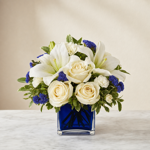 Sapphire Elegance Standard – compact arrangement of creamy white roses, white Asiatic lilies, mini carnations and blue statice in a cobalt blue cube vase, smaller lighter version